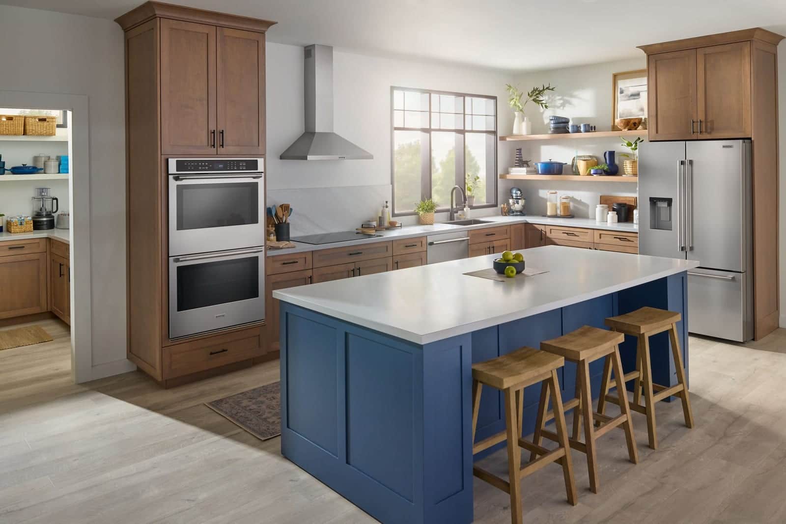 A wide kitchen with a stainless Maytag double wall oven set into wood cabinetry, a blue island with three wood stools, and a stainless French-door refrigerator at the right