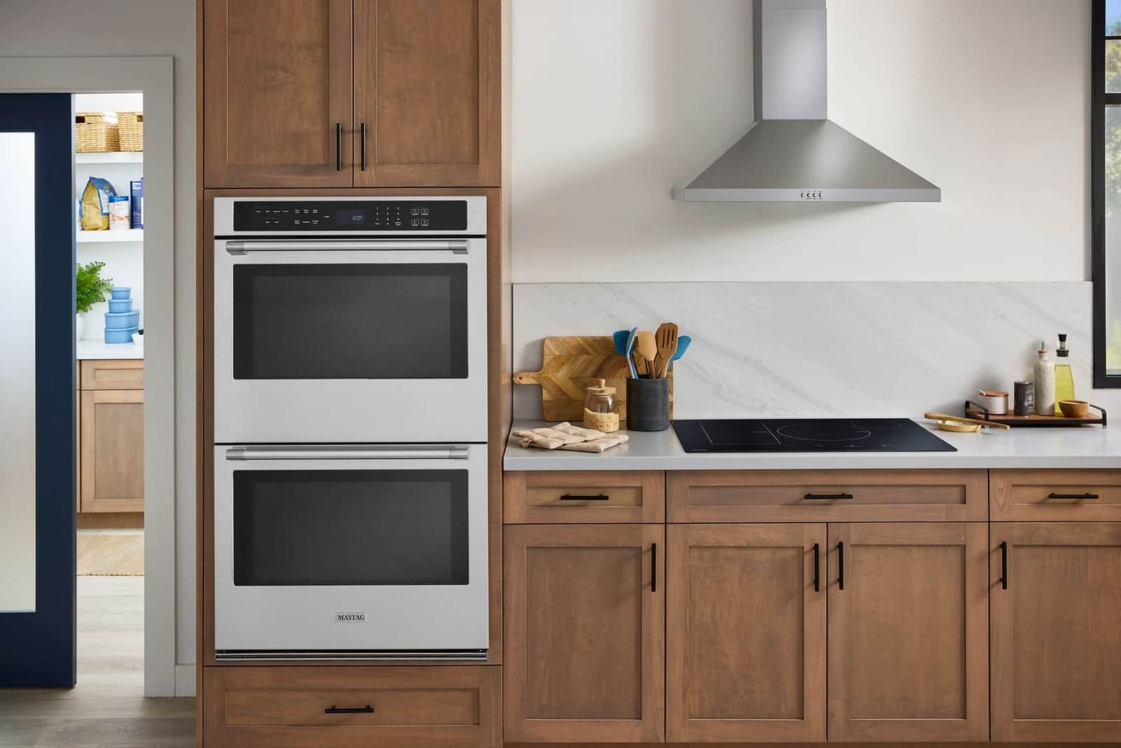 A stainless Maytag double wall oven in wood cabinetry beside a white counter with a black induction cooktop under a stainless chimney hood