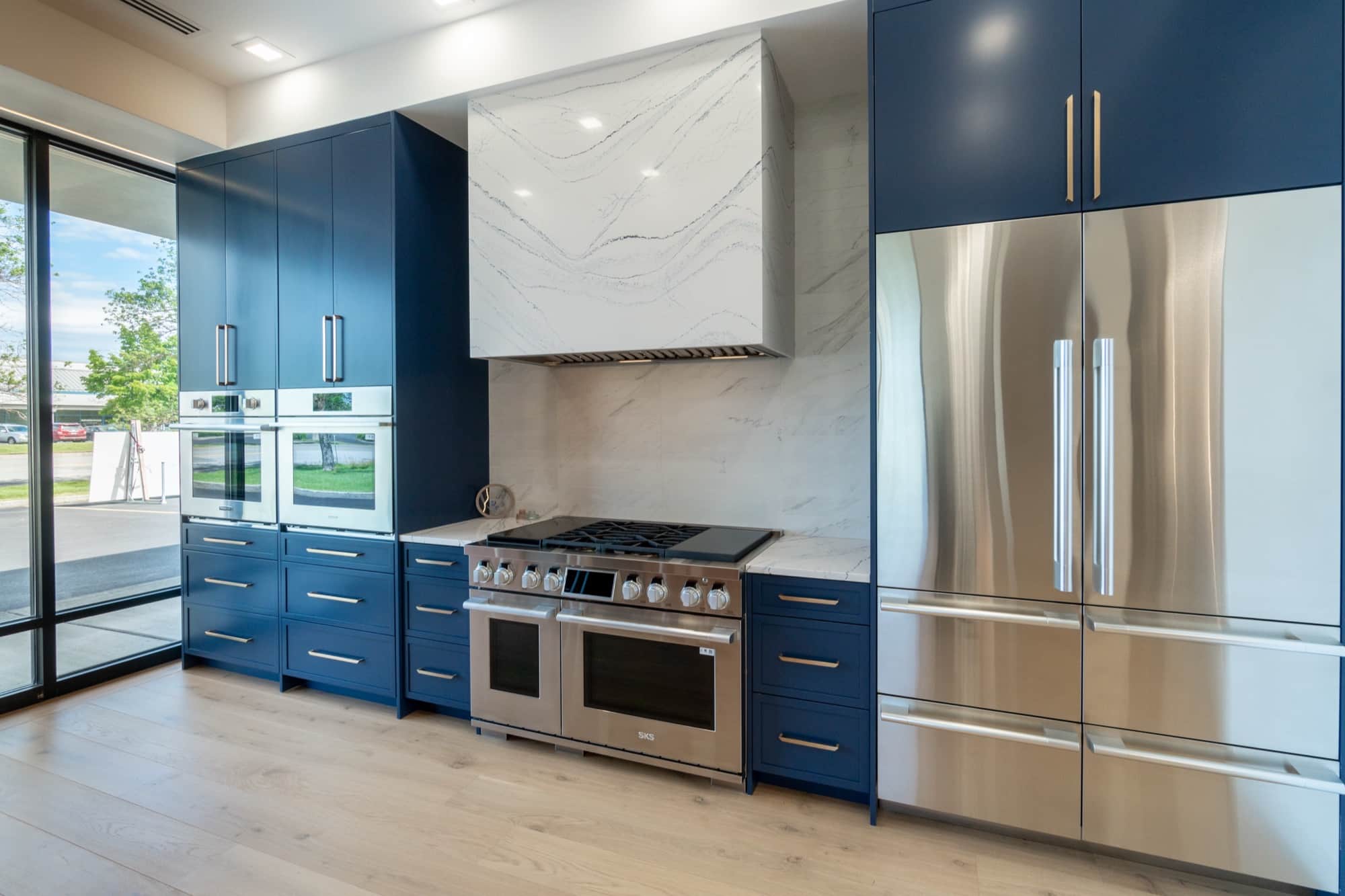 A kitchen vignette with navy cabinetry, two stainless wall ovens, a professional dual-oven range with a griddle under a white veined slab chimney hood, and a built-in stainless French-door refrigerator