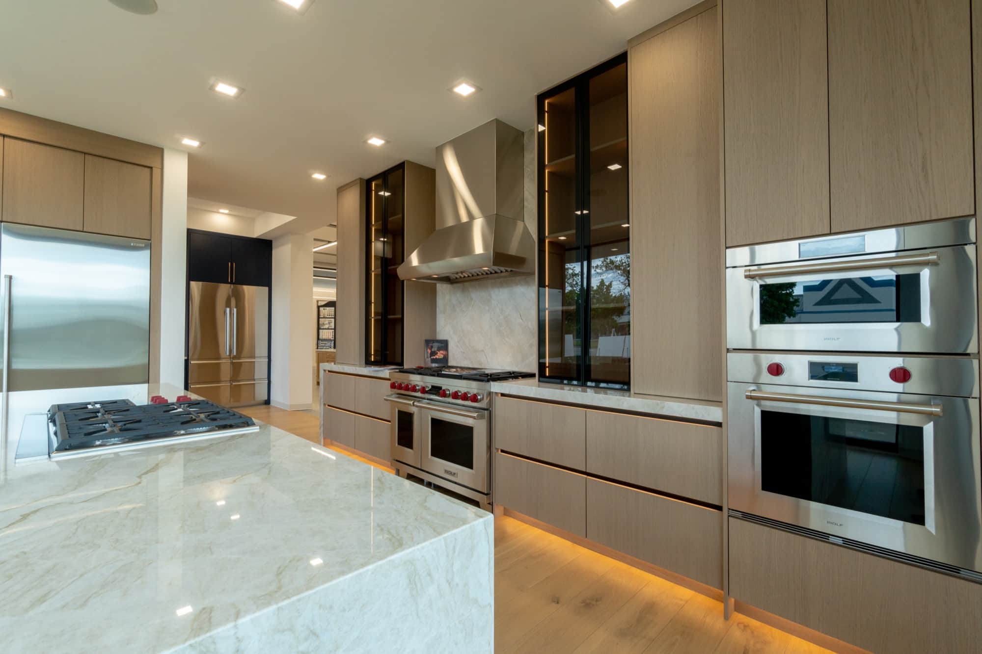 A display kitchen on the Post Falls showroom floor seen down its length, with a Wolf professional range under a stainless chimney hood, a Wolf wall oven and speed oven column at right, and refrigeration along the left wall